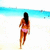 A woman in a bikini walks along a beach with blue water and white sand, viewed from behind.