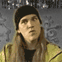 A person with long brown hair and a black beanie wears a yellow jacket over a black shirt against a gray patterned background with white swirls.