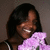 A person with dark hair holds a large purple flower, smiling broadly while wearing a sleeveless top.