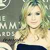 A woman with wavy blonde hair wears a dark strapless dress against a gray backdrop with partial text reading 'THE GRAMMY AWARDS' and a circular logo.