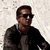 A man with short dark hair and sunglasses wears a dark jacket over a light shirt against a textured light background.