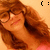 A pixelated image of a person with long brown hair and glasses, wearing a pink top against an orange background.