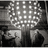 Pixelated grayscale image of a person holding a large circular light fixture with multiple bulbs, accompanied by cyan text elements on black background.