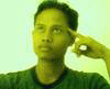 A person in a green shirt with a dark collar makes a finger gesture near their head against a light background.