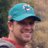 Pixelated static image of a person wearing a teal and dark blue cap, red shirt, and dark sunglasses, with a blurred background.