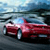 A red sports car with a white roof is positioned on a road, viewed from a low angle with a blurred background of sky and water.