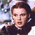Pixelated image of a person with dark hair wearing a dark jacket over a white shirt, holding a small object near their face against a blurred background.