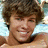A pixelated image of a person with curly brown hair smiling, wearing a dark collar, against a light blue background with a pink symbol in the corner.