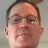 Pixelated close-up of a person's face with short dark hair, wearing glasses, against a light gray background.