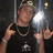 Pixelated image of a person wearing dark clothing and multiple necklaces, holding up two fingers in a gesture against a dark background with light reflections.