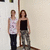 Two women stand side-by-side against a light wall; one wears a patterned sleeveless top and dark pants, the other a black sleeveless top and camouflage pants.