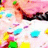 Pixelated abstract composition with scattered colored blocks in pink, blue, yellow, green, and purple hues against a light background.