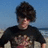 Pixelated image of a person with dark curly hair wearing a black shirt, standing against a beach background with blue sky and water.