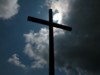 Silhouette of a cross against a cloudy sky with sunlight breaking through, rendered in low-resolution pixels.