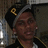 A person wearing a black cap with yellow 'P' and gray trim, dark jacket with gold stripes, white shirt, and small earpiece in right ear.