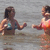 Two people in swimwear stand in shallow water, one with a blue bikini top and the other with an orange swimsuit, both facing left.