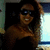 A pixelated image of a person with long dark hair wearing dark sunglasses and a dark top, smiling with one arm raised near their head against a dark background with light areas on the right.