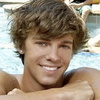 A static image of a person with light brown curly hair resting their arms on a surface near a pool, wearing no shirt.