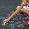 A person's arm and hand draw a yellow heart on a dark surface with colorful chalk hearts scattered around.