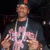 A person wearing a dark shirt with pink text, silver chain necklace, and red-topped cap making hand gestures against a dark background.
