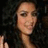 A woman with long dark curly hair and light makeup smiles slightly, her right hand raised in a wave against a dark background.