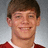 Pixelated portrait of a person with short brown hair wearing a red shirt with white collar, smiling against light gray background.