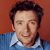 Pixelated portrait of a person with dark hair wearing a blue striped shirt over a white collared shirt against a solid reddish-brown background.