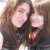 Two women with long brown hair pose closely together, one wearing a red necklace, against a light background.