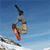 Pixelated static image of a skier in mid-air against blue sky background with snow-covered ground below, wearing gray jacket and beige pants, red ski boots visible, black helmet or headgear on top.