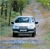 Pixelated image of a light blue van with dark blue accents parked on a paved surface, surrounded by green foliage and trees.