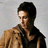 A person with dark hair and glasses wears a brown jacket over a black shirt against a light gray background.