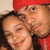 Two people in close proximity, one wearing a red cap and dark clothing, the other with long dark hair, both smiling against a light background.