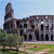 Pixelated static image of a large building with a tall tower, dark windows, and a grassy foreground under a light blue sky.