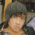 Pixelated close-up of a person with light skin wearing a dark gray beanie, brown hair visible beneath the hat, and a yellowish-brown garment in background.