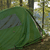 Green tent with dark straps in a forest setting, surrounded by trees and foliage under daylight.
