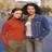 Pixelated static image of two people standing close together, one in red top and green pants, the other in blue jacket over white shirt with dark hair.
