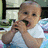 Pixelated image of a baby in a white onesie with arms raised, mouth open, against a blurred indoor background with green and blue tones.