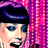 A close-up of a person with dark hair and heavy eye makeup against a pink and purple pixelated background.