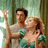 Two people in a close embrace, one holding the other's hand up while both wear light-colored tops against a blurred green background.