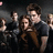 A group of people in dark clothing pose together in a dimly lit scene with a gray background, some raising hands.