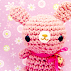 Pink knitted bunny with black eyes and golden nose, wearing a pink ribbon with a small golden bell, set against a pink background with white and yellow floral patterns.