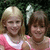 Two girls in pink and white tops with a large white bow, standing against green foliage background.