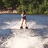 A person stands on a watercraft in motion, creating white spray against blue water with green trees behind.