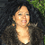 Pixelated image of a person with light skin tone, dark hair, and a dark jacket, smiling with eyes closed against a dark background with green foliage.