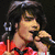 A pixelated image shows a person with dark hair holding a microphone, wearing red clothing against a black background.
