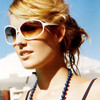 A person with light brown hair tied back wears white-framed sunglasses and a blue beaded necklace against a bright blue sky background.