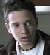 Static pixelated image of a person in a white shirt and dark jacket against a blurred background with visible dithering and compression artifacts.