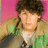 A person with curly brown hair wears a green jacket with a beige collar over a white shirt against a pink background.