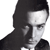 Pixelated grayscale portrait of a person with long dark hair, wearing a dark jacket over a light shirt, facing slightly right with a neutral expression.