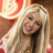 A blonde woman with long hair smiles in a of four , wearing a white top with red accents and a dark necklace, against a red and orange circular background